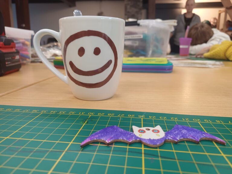 Selbst gebastelte Fledermaus und eine Tasse mit Smiley auf einer Bastelmatte